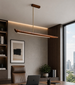 A modern linear pendant light with a warm wood-grain finish and a slim brass rod hangs over a office desk.