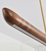 Close-up of a modern linear light fixture with a curved, rounded wooden shade in a warm medium-brown finish, showing a white diffuser strip and a gold-toned hanging rod.