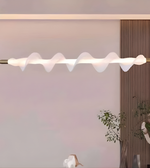Modern ceiling light fixture with wavy design in a room with a plant and wooden panel.