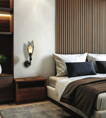 A modern wall-mounted sconce with a black metal frame and a warm glowing glass shade, mounted on a white wall in a contemporary bedroom next to a wooden nightstand and bed.