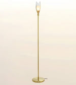 A modern floor lamp with a slim brass-colored metal pole, a round base, and a clear tulip-shaped glass shade that glows with a warm light, set against a soft beige gradient background.