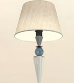 A modern table lamp with a pleated beige fabric drum shade, a light blue glass sphere, and sculptural white and gray geometric bases on a warm off-white background.