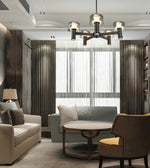 A modern living room interior featuring a multi-light chandelier with cylindrical glass shades, a light gray sofa, a cream sofa, a round wooden coffee table, and a brown armchair with a yellow cushion, set against dark wood paneling and sheer curtains.