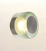 A modern cylindrical wall light fixture with a clear glass shade, a silver metallic side, and a circular gray backplate, emitting a warm yellow light against a plain off-white background.