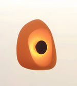 A sculptural, organic-shaped light fixture or lamp with a warm orange-brown outer shell and a glowing yellow-orange center, set against a plain off-white background.