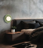 A modern bedroom scene featuring a decorative green sunburst wall mirror mounted on a textured gray concrete wall above a dark wood nightstand and bed.