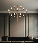 A modern multi-light chandelier with a brushed brass/gold metal frame and frosted white globe bulbs, hanging in a styled living room with a dark gray wall, decorative molding, and a dark sofa below.