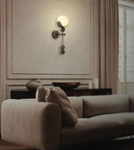 A modern wall-mounted sconce with a frosted round globe and dark metal accents, mounted on a beige paneled wall above a beige sofa in a styled living room.