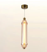 A modern cylindrical pendant light with a warm glowing glass shade and antique brass metal accents, suspended against a soft beige gradient background.