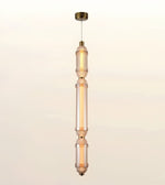 A modern vertical pendant light fixture with three stacked cylindrical glass shades in a warm frosted amber tone, accented by brass-toned metal hardware and a matching ceiling canopy, shown against a plain off-white background.
