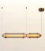 A modern linear pendant light fixture with two cylindrical glass shades and textured metallic end caps, suspended by thin cables from a round ceiling canopy against a plain off-white background.