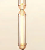 A close-up of a cylindrical pendant light fixture with a warm white glowing bulb inside a clear glass shade, accented by gold-toned metal bands and textured details on a light beige background.