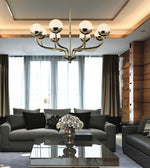 A modern multi-arm chandelier with spherical white glass globes and a brushed brass finish hangs in a contemporary living room with a gray sofa, glass coffee table, and large windows.
