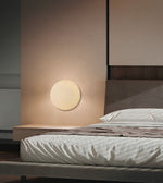 A modern minimalist bedroom featuring a round, flush-mounted warm white wall sconce on a beige wall, next to a low wooden nightstand and a bed with white textured bedding.