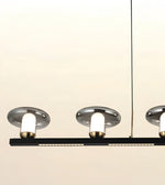 Modern linear pendant light fixture with three illuminated cylindrical bulbs in brushed silver shades and a black horizontal bar, shown against a plain light beige background.