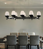 A modern 7-light linear chandelier with textured white glass drum shades and black metal arms, suspended over a dining table with gray chairs in a dining room.