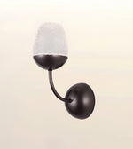 A modern wall-mounted sconce with a dark bronze-finished metal body, featuring a curved arm and a textured frosted glass shade, shown against a light beige background.