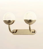 A modern double-light wall sconce with a polished brass or gold-finished metal frame, featuring two spherical textured white glass shades mounted on a horizontal bar.
