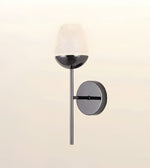 Modern wall-mounted sconce with a cylindrical textured white shade, a matte black bowl, a slim black vertical stem, and a round gray wall plate on a warm off-white background.