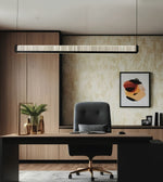 A modern office interior featuring a long rectangular linear pendant light hanging above a desk with a black office chair. The room has light wood paneling, beige patterned wallpaper, framed abstract wall art, and a gray rug.