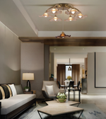 A modern living room interior featuring a decorative ceiling light fixture with multiple warm glowing bulbs and flower-like shades, along with a sofa, armchair, coffee table, and dining area in the background.