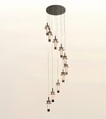 A modern multi-light linear pendant chandelier with nine cascading cylindrical glass shades and dark metal hardware, suspended from a circular ceiling canopy against a light beige background.