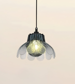 A hanging pendant light with a black cord, a dark textured central shade, and translucent petal-like outer shades around a glowing frosted bulb, shown against a plain light beige background.