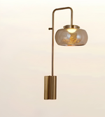 A modern wall sconce lamp with a brushed brass/gold metal frame, featuring a curved arm, a cylindrical wall mount, and a frosted glass shade illuminated from within.