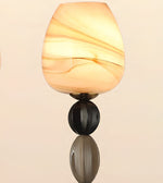 A modern wall sconce featuring a warm, glowing teardrop-shaped glass shade with cream and amber swirls, mounted on a dark metallic stem with a reflective base against a light beige background.