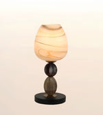 A modern table lamp with a rounded, teardrop-shaped shade made of marbled amber and cream glass, supported by a dark brown glass stem and a round black base, set against a soft beige gradient background.