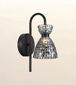 A modern wall sconce light fixture with a matte black curved arm and circular wall mount, featuring a clear glass shade made of reflective mirrored tiles that reflect warm light.