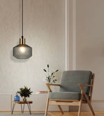 A modern pendant light with a textured dark gray glass shade and a brass-colored collar, hanging in a minimalist room next to a gray upholstered wooden armchair.