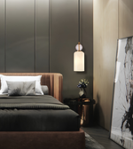 A modern bedroom interior featuring a bed dressed in dark charcoal bedding and white pillows, with a warm brown upholstered headboard and footboard. A black pendant light hangs above a small side table with a vase of flowers, and a large framed abstract print leans against the wall on the right.