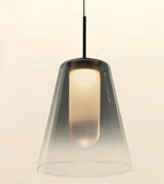 A modern pendant light with a tapered clear glass shade and a black hanging cord, shown against a plain light beige background.