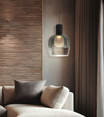 A modern pendant light with a clear glass dome shade and a black cylindrical top, hanging above a beige sofa in a styled living room with wood paneling and curtains.