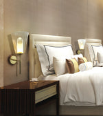 A modern bedroom scene featuring a bed dressed in white linens with decorative pillows, a dark wood nightstand, and two illuminated wall sconces mounted on a beige textured wall.