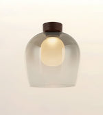 A modern pendant light with a rounded, dome-shaped translucent smoky glass shade, a warm glowing bulb inside, and a dark brown cylindrical cap at the top, shown against a plain off-white background.