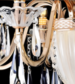 Close-up detail of a modern chandelier with a polished gold-toned metal frame, white translucent glass shades, and hanging clear crystal prisms against a black background.