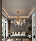 An ornate multi-arm chandelier with a gold-toned metal frame and clear crystal droplets, hanging in a modern living room with a gray sofa and dark gray paneled walls.