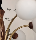 Close-up of a modern multi-arm chandelier with frosted white glass globe shades, curved brass metal arms, and dark brown finials, with text labels pointing to the glass and metal materials.