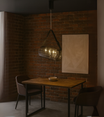 Cylindrical glass pendant light with strap suspension hanging above a dining table in a brick-walled room.