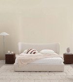 Modern beige upholstered bed with neutral bedding, wood nightstands, floor lamp, and plush rug