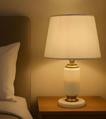 Table lamp with a white shade and gold base on a wooden nightstand next to a bed.
