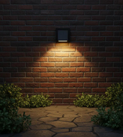 Wall-mounted outdoor light fixture on a brick wall with a stone pathway and greenery.