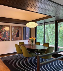 Dining room with round table and green chairs, large windows showing trees, and colorful wall art.