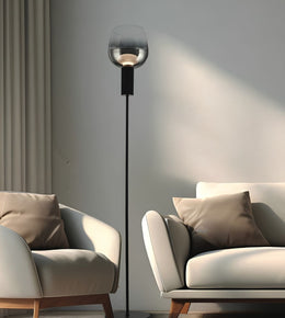 A modern black floor lamp with a clear glass globe shade stands between two upholstered armchairs in a minimalist living room setting.