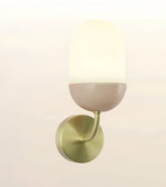 A modern wall-mounted sconce with a round brushed brass base, a curved arm, and a frosted white dome-shaped shade that glows warm white when lit, shown against a plain light background.