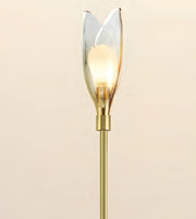 A modern tulip-shaped wall sconce with a warm glowing light bulb inside a clear glass petal-shaped shade, mounted on a brushed gold metal stem against a solid beige background.