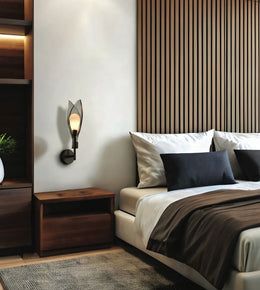 A modern wall-mounted sconce with a black metal frame and a warm glowing glass shade, mounted on a white wall in a contemporary bedroom next to a wooden nightstand and bed.