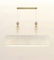 A modern rectangular pendant light fixture with a large white textured drum shade, a brushed brass canopy, and two hanging rose gold beads.